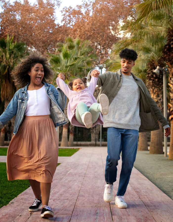 Sonder A joyful family walks together in a park, swinging a child between them amidst palm trees and autumn scenery.